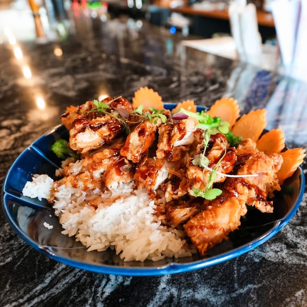 Crispy fried chicken with sauce, white rice, and carrot garnishes in a blue bowl on a dark counter.