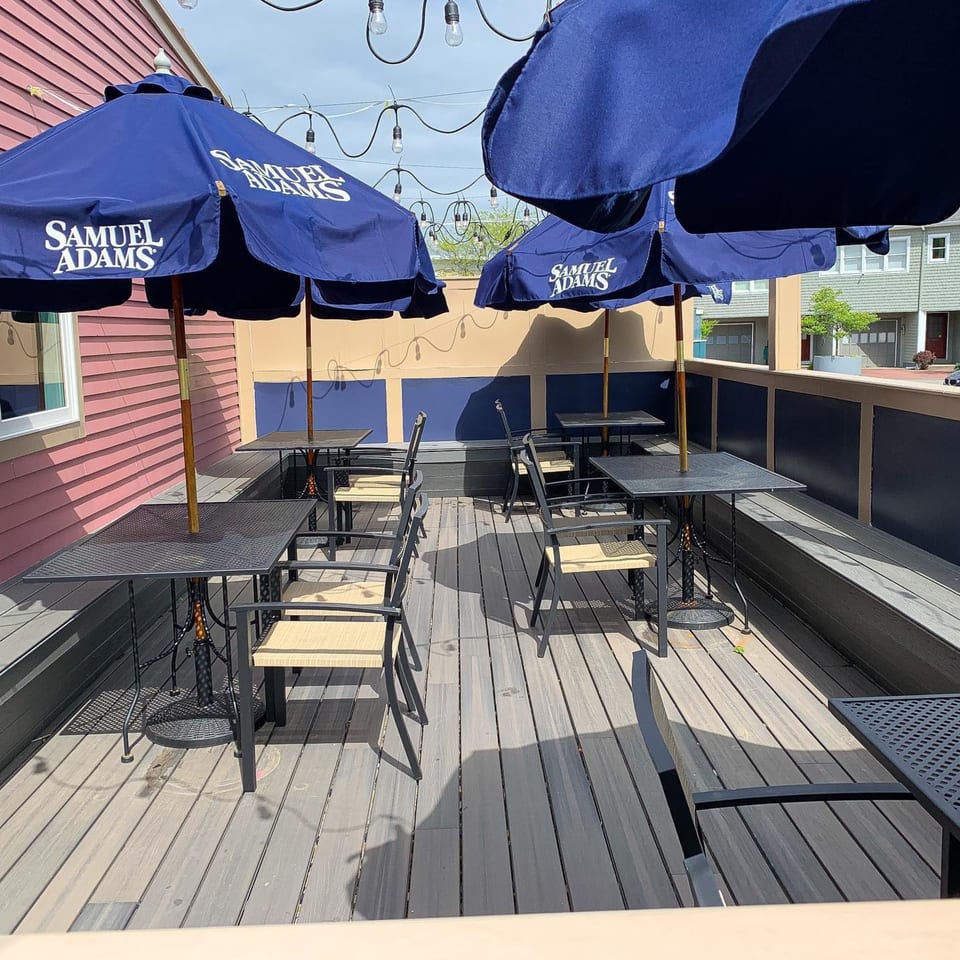 Outdoor patio with black metal tables, light chairs, and blue Samuel Adams umbrellas under string lights.