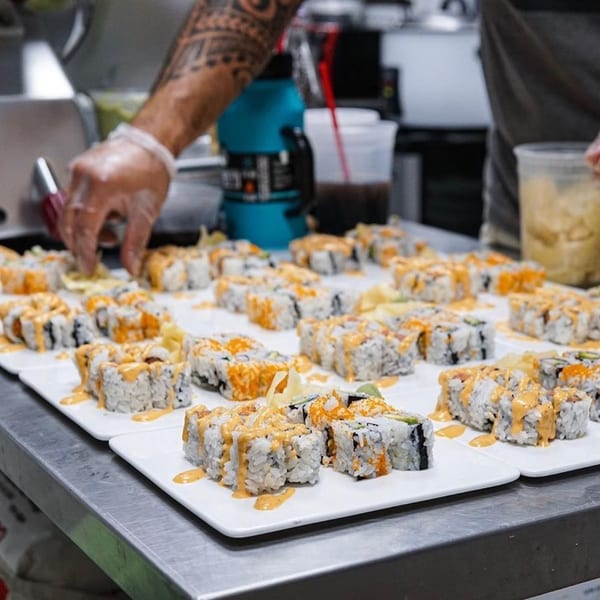 A chef's tattooed arm and gloved hand arranging many plates of sushi rolls with orange sauce.