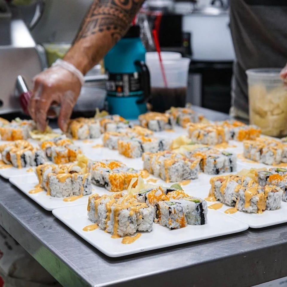 A chef's tattooed arm and gloved hand arranging many plates of sushi rolls with orange sauce.