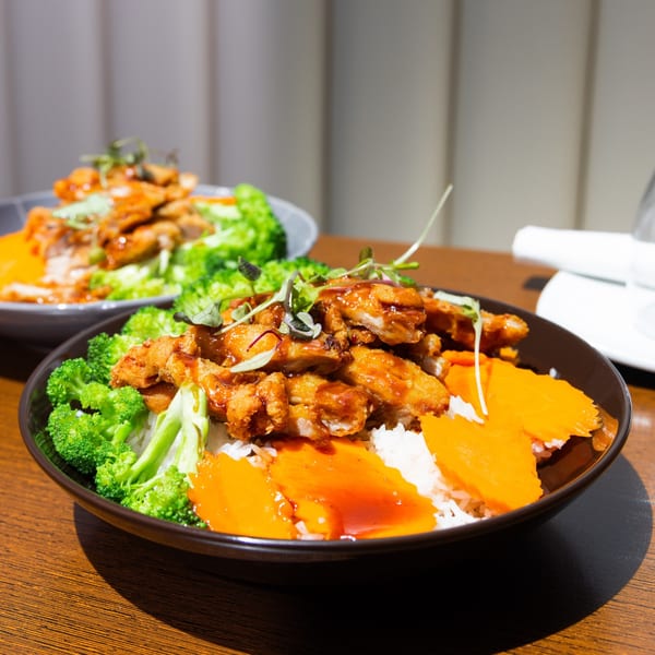 Two bowls of Bleu Ginger's Asian-fusion chicken, rice, broccoli, and carrots on a wooden table.