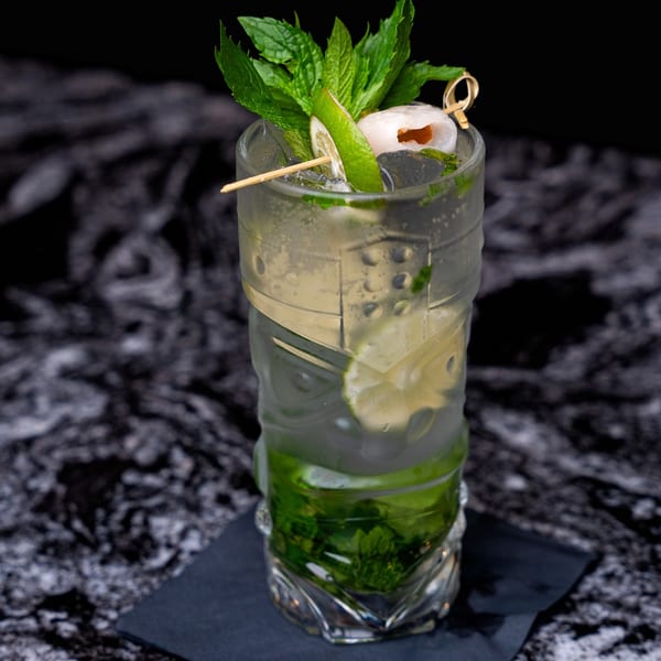 A tall, clear glass of a mojito cocktail with mint, lime, and a lychee garnish.