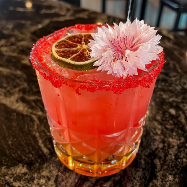 A vibrant red cocktail with a sugared rim, garnished with a dried lime slice and a pink flower.